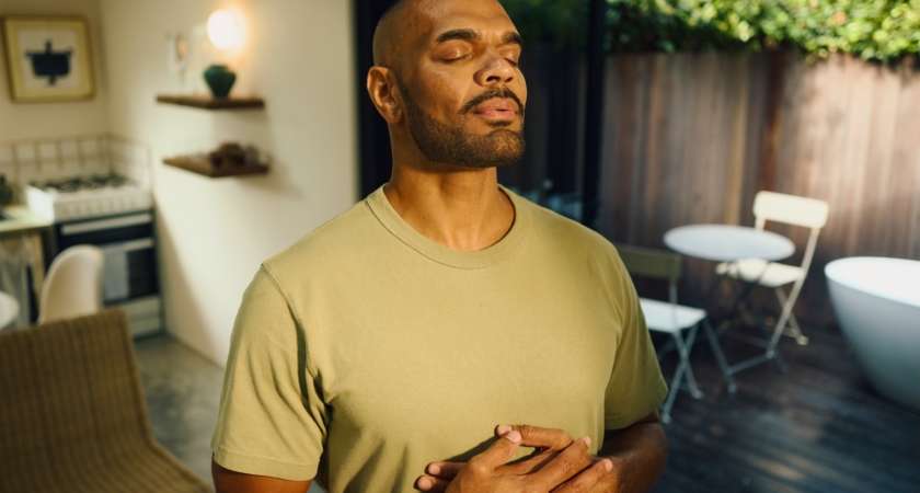 Serene young aboriginal australian man practicing mindfulness, taking deep breaths with closed eyes and hands on chest, enjoying a peaceful morning at home